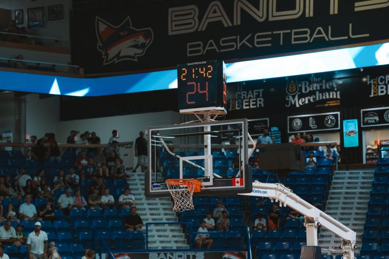 Close-up of a basketball hoop inside a stadium with a scoreboard and audience in the background.