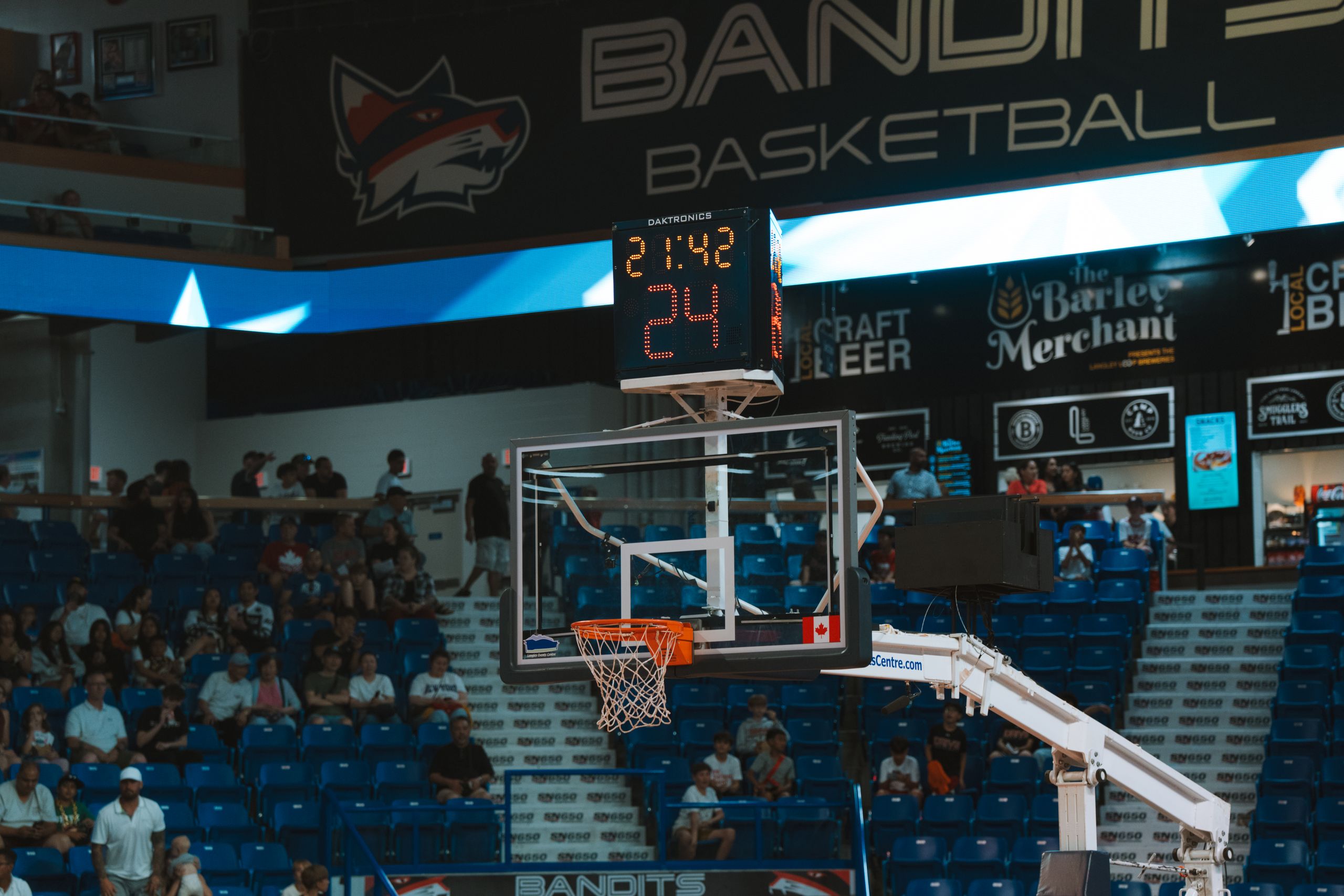 Close-up of a basketball hoop inside a stadium with a scoreboard and audience in the background.